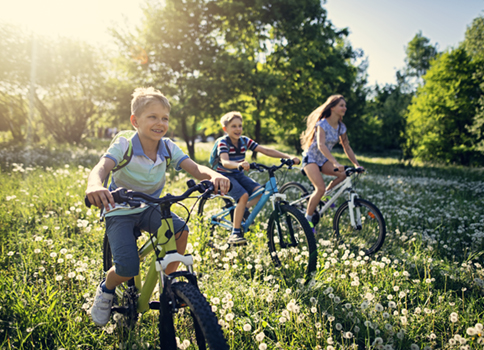 Two boys and a girl ride bikes through a field on a pretty day