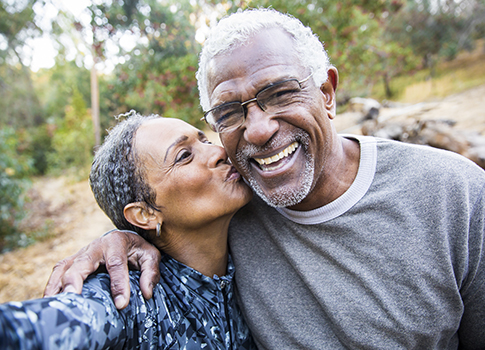 Older man in an outdoor setting smiles as wife kisses him on the cheek