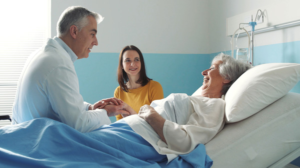 Physician sits in a hospital bed next to an elderly woman patient, smiling and holding her hand, while younger family member looks on. 