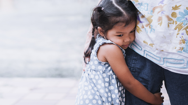 A young girl hugs the leg of her parent.