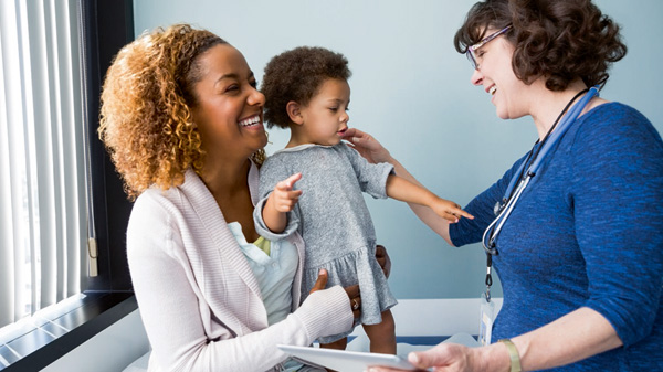 In a welcoming doctor office setting, a mother joyfully holds up her toddler daughter while a healthcare professional conducts an exam.