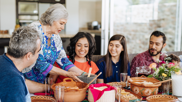 Grandmother places dish on table with lots of other food, and with family and friends gathered around.