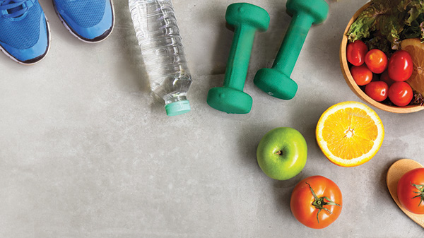 Overhead view of a bowl of fruits and vegetables, hand weights, water bottle, and a pair of running shoes.