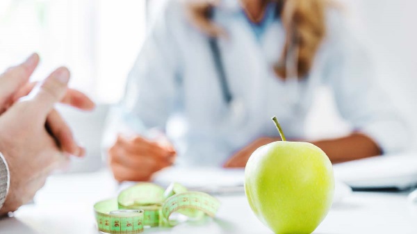 An apple and a tape measure in foreground on a table between two people talking.