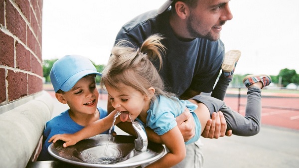 Father helps daughter drink from a water fountain while her brother waits for his turn
