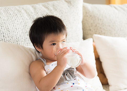 A baby sitting a couch and drinking a glass of milk