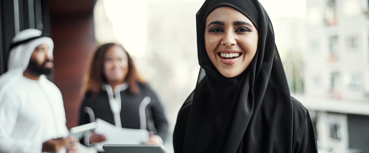 A business woman in hijab holds a tablet and smiles at the camera