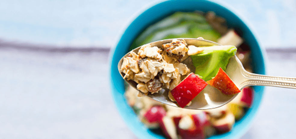 A spoon holding granola and apples over a bowl