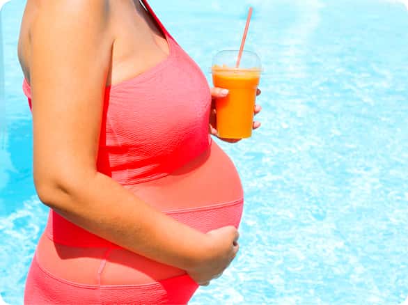 אישה בהריון בבריכה מחזיקה כוס מיץ תפוזים
