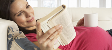 אישה הרה נחה על הספה, שותה משקה חם וקוראת ספר