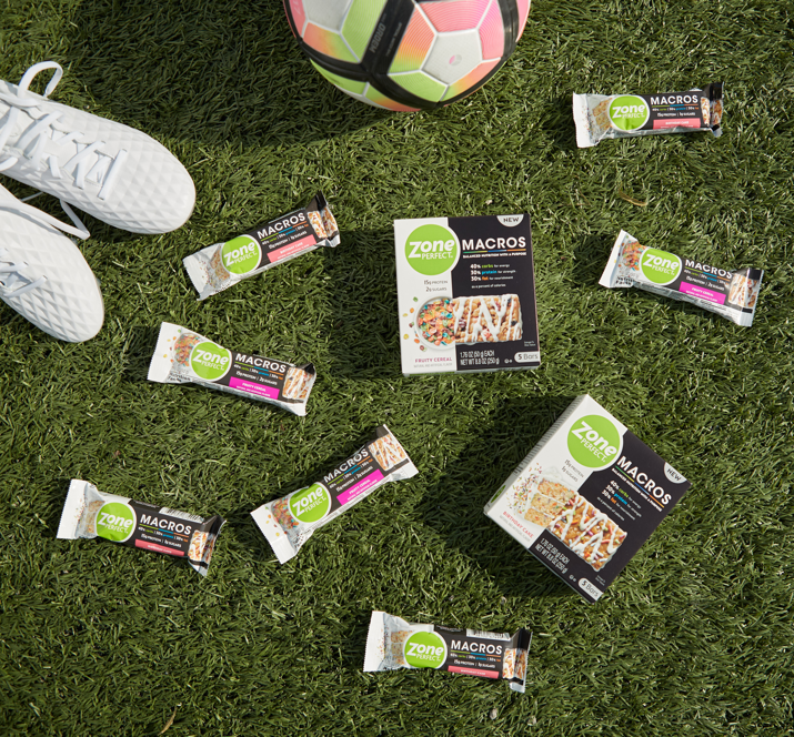 Soccer cleats, soccer ball, and ZonePerfect Macros bars sitting on a soccer field